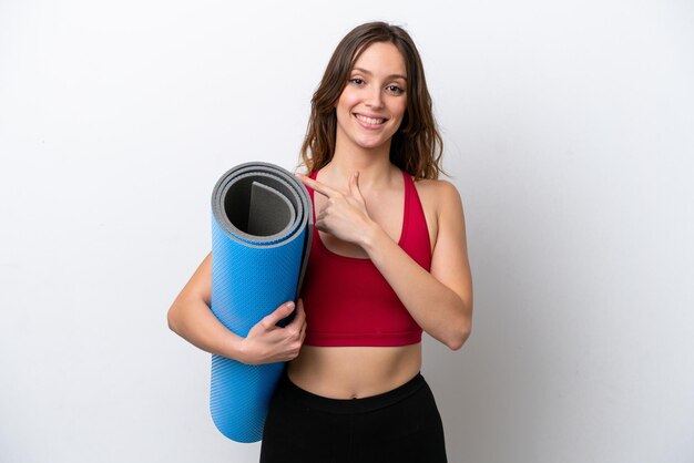 Jeune femme caucasienne sportive allant à des cours de yoga tout en tenant un tapis isolé sur fond blanc pointant vers le côté pour présenter un produit