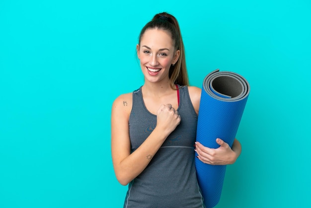 Jeune femme caucasienne sportive allant à des cours de yoga tout en tenant un tapis isolé sur fond bleu célébrant une victoire