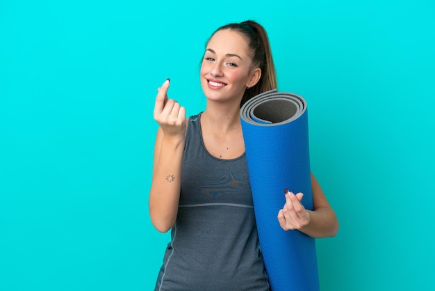 Jeune femme caucasienne sportive allant à des cours de yoga tout en tenant un tapis isolé sur fond bleu faisant un geste d'argent