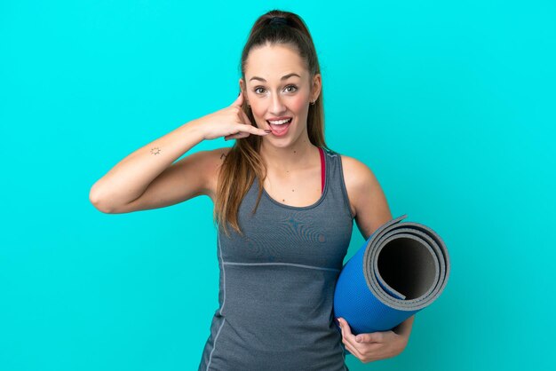 Jeune femme caucasienne sportive allant à des cours de yoga tout en tenant un tapis isolé sur fond bleu faisant un geste de téléphone Appelez-moi signe de retour
