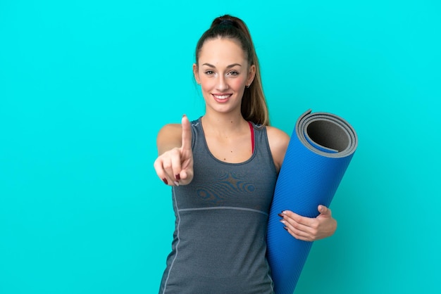 Jeune femme caucasienne sportive allant à des cours de yoga tout en tenant un tapis isolé sur fond bleu montrant et levant un doigt