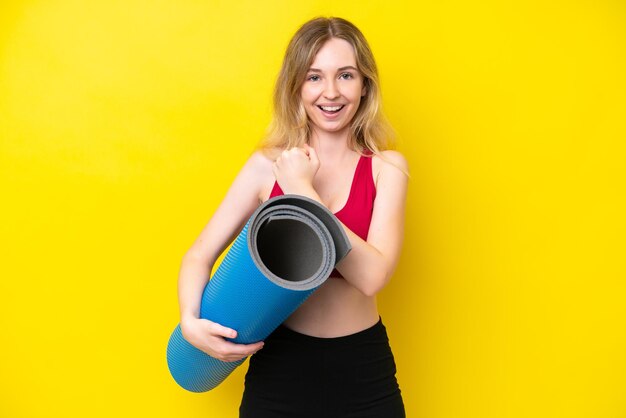 Jeune femme caucasienne sportive allant à des cours de yoga tout en tenant un tapis isolé sur fond jaune célébrant une victoire