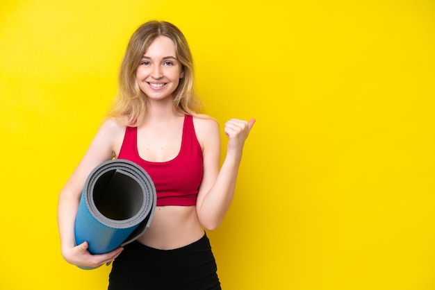 Jeune femme caucasienne sportive allant à des cours de yoga tout en tenant un tapis isolé sur fond jaune pointant vers le côté pour présenter un produit