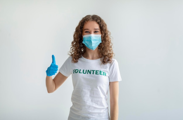 Jeune femme caucasienne en t-shirt bénévole blanc montrant le pouce vers le haut portant un masque médical et des gants