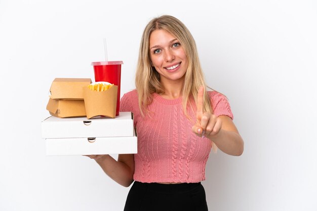 Jeune femme caucasienne tenant des aliments gras isolés sur fond bleu montrant et levant un doigt