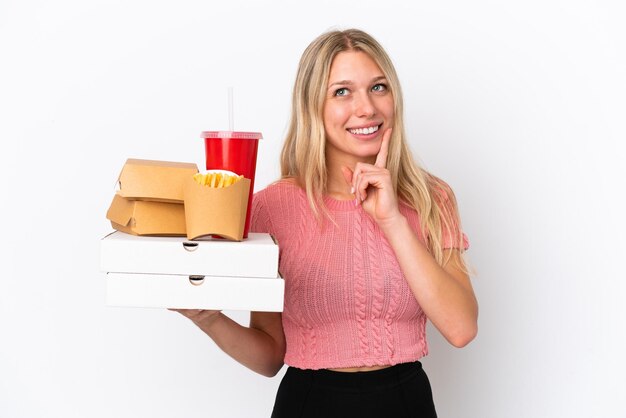 Jeune femme caucasienne tenant des aliments gras isolés sur fond bleu en pensant à une idée tout en levant les yeux