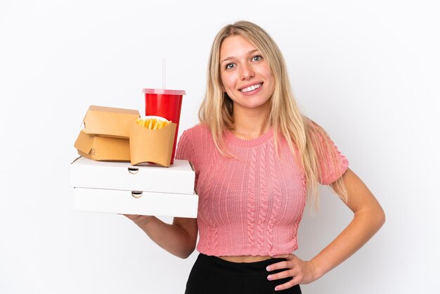 Jeune femme caucasienne tenant des aliments gras isolés sur fond bleu posant avec les bras à la hanche et souriant