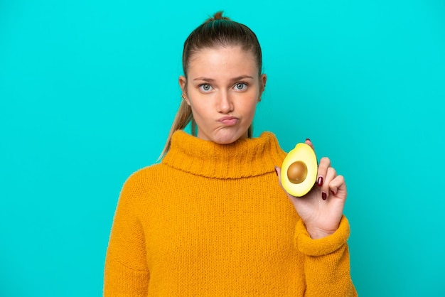 Jeune femme caucasienne tenant un avocat isolé sur fond bleu avec une expression triste