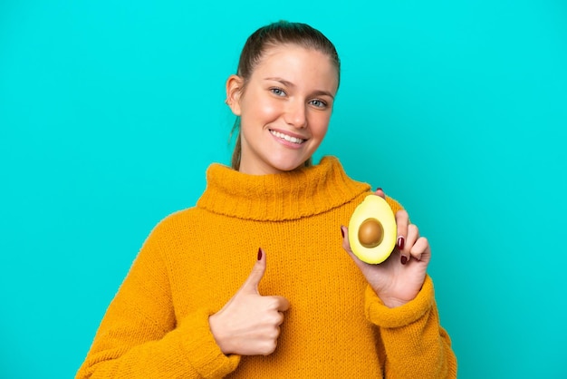 Jeune femme caucasienne tenant un avocat isolé sur fond bleu avec les pouces vers le haut parce que quelque chose de bien s'est produit