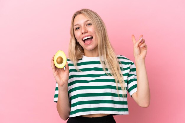 Jeune femme caucasienne tenant un avocat isolé sur fond rose souriant et montrant le signe de la victoire