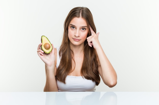 Jeune femme caucasienne tenant un avocat pointant son temple avec le doigt, pensant, concentré sur une tâche.