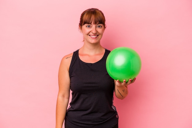 Jeune femme caucasienne tenant le ballon pour faire du sport isolé sur fond rose heureux, souriant et joyeux.