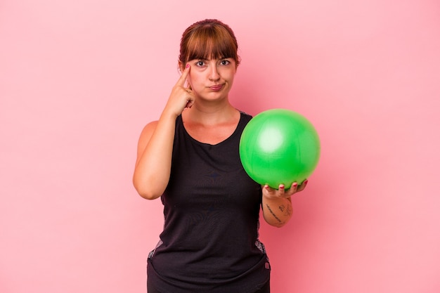 Jeune femme caucasienne tenant le ballon pour faire du sport isolé sur fond rose pointant le temple avec le doigt, pensant, concentré sur une tâche.