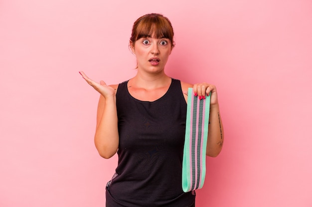 Jeune femme caucasienne tenant des bandes élastiques isolées sur fond rose surprise et choquée.