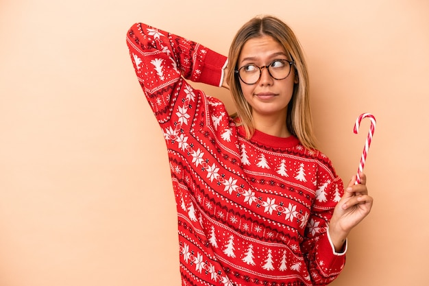 Jeune femme caucasienne tenant un bâton de Noël isolé sur fond beige touchant l'arrière de la tête, pensant et faisant un choix.