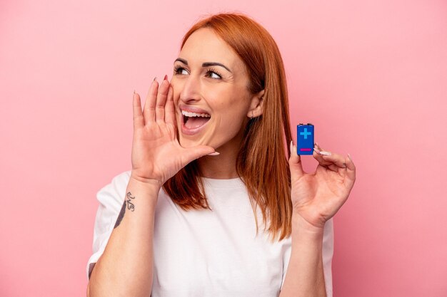 Jeune femme caucasienne tenant une batterie isolée sur fond rose criant et tenant la paume près de la bouche ouverte.