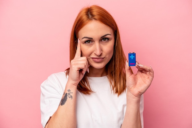 Jeune femme caucasienne tenant une batterie isolée sur fond rose pointant le temple avec le doigt, pensant, concentrée sur une tâche.