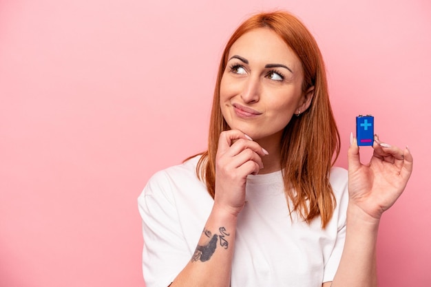 Jeune femme caucasienne tenant une batterie isolée sur fond rose regardant de côté avec une expression douteuse et sceptique