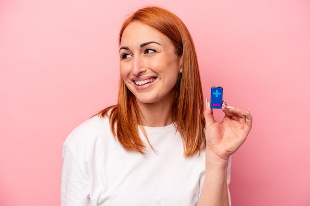 Jeune femme caucasienne tenant une batterie isolée sur fond rose regarde de côté souriant gai et agréable