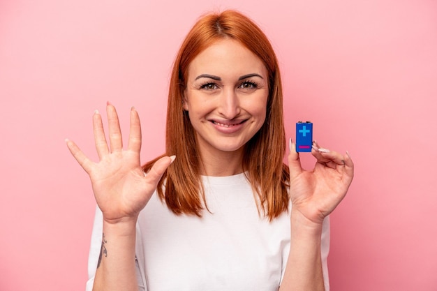 Jeune femme caucasienne tenant une batterie isolée sur fond rose souriant joyeux montrant le numéro cinq avec les doigts.