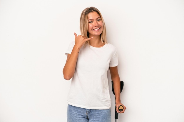 Jeune femme caucasienne tenant une béquille isolée sur fond blanc montrant un geste d'appel de téléphone portable avec les doigts.
