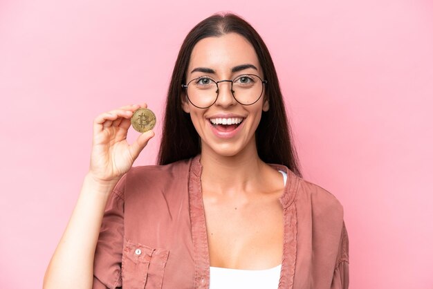 Jeune femme caucasienne tenant un Bitcoin isolé sur fond rose avec une expression faciale surprise et choquée