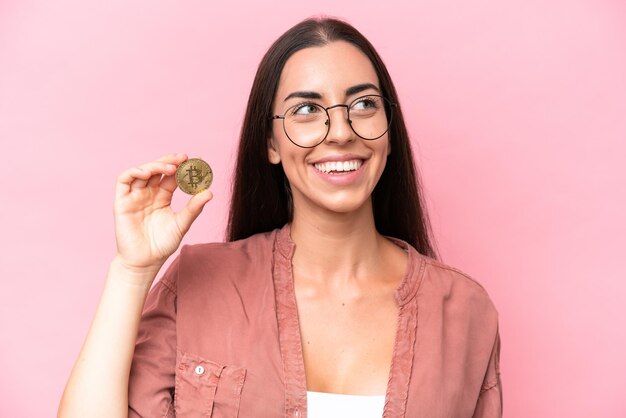 Jeune femme caucasienne tenant un Bitcoin isolé sur fond rose levant les yeux en souriant