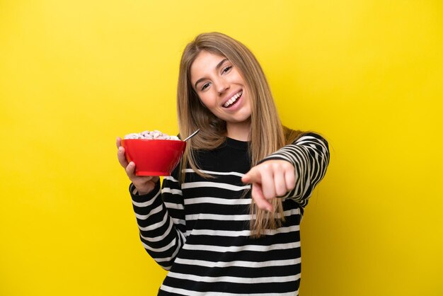 Jeune femme caucasienne tenant un bol de céréales isolé sur fond jaune pointe le doigt vers vous avec une expression confiante