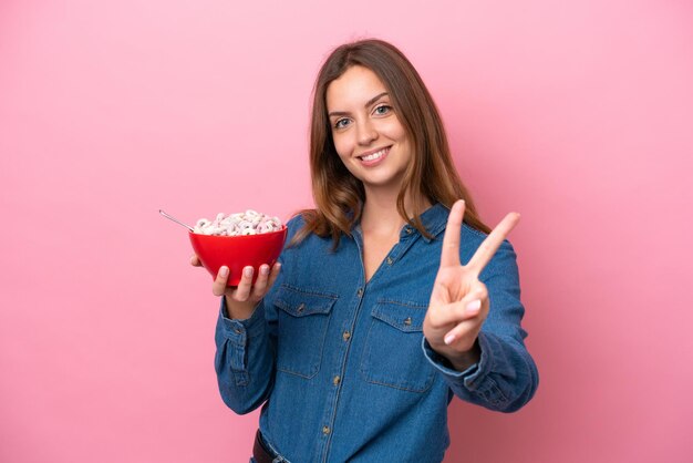 Jeune femme caucasienne tenant un bol de céréales isolé sur fond rose souriant et montrant le signe de la victoire