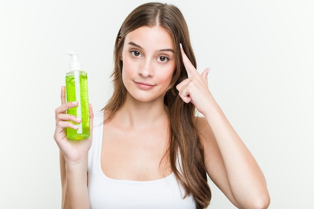 Jeune femme caucasienne tenant une bouteille d'aloe vera pointant sa tempe avec le doigt, pensant, concentrée sur une tâche.