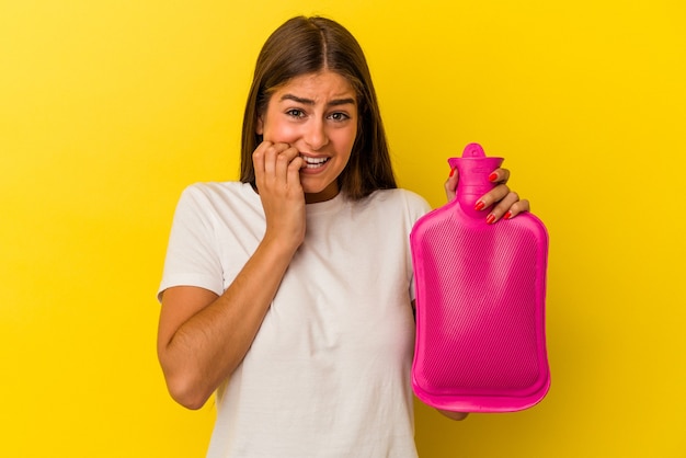 Jeune femme caucasienne tenant une bouteille d'eau chaude isolée sur fond jaune se rongeant les ongles, nerveuse et très anxieuse.