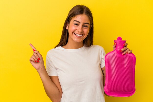 Jeune femme caucasienne tenant une bouteille d'eau chaude isolée sur fond jaune souriant et pointant de côté, montrant quelque chose dans un espace vide.
