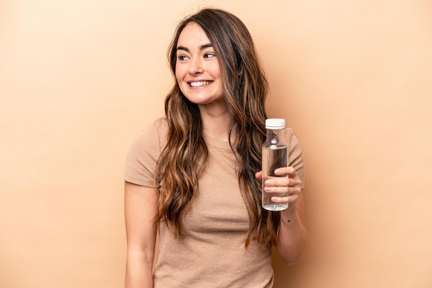 Jeune femme caucasienne tenant une bouteille d'eau isolée sur fond beige regarde de côté souriant gai et agréable