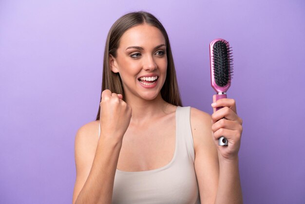 Jeune femme caucasienne tenant une brosse à cheveux isolée sur fond violet célébrant une victoire