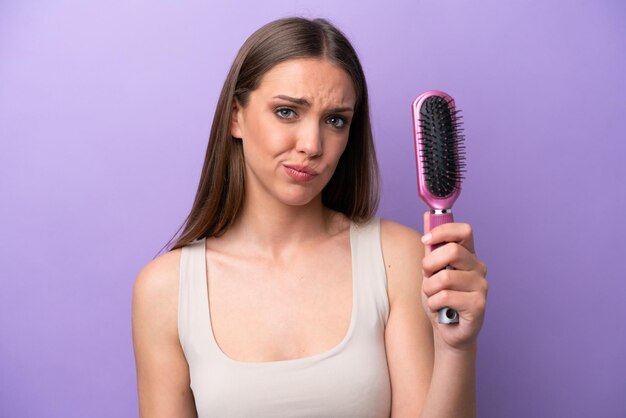 Jeune femme caucasienne tenant une brosse à cheveux isolée sur fond violet avec une expression triste