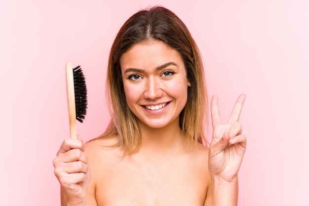 Jeune femme caucasienne tenant une brosse à cheveux montrant le signe de la victoire et souriant largement.