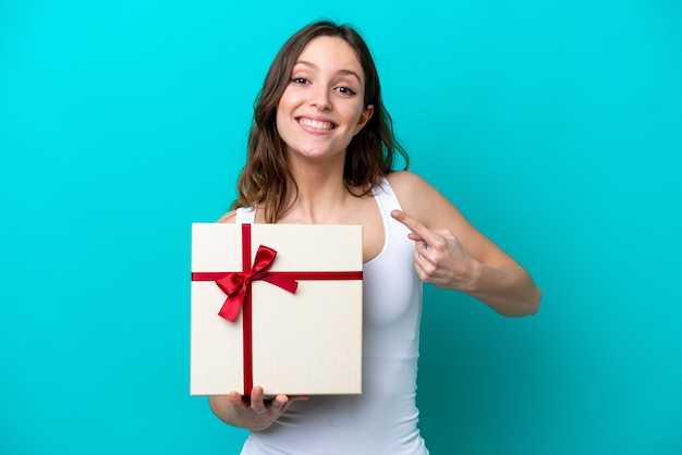 Jeune femme caucasienne tenant un cadeau isolé sur fond bleu avec une expression faciale surprise