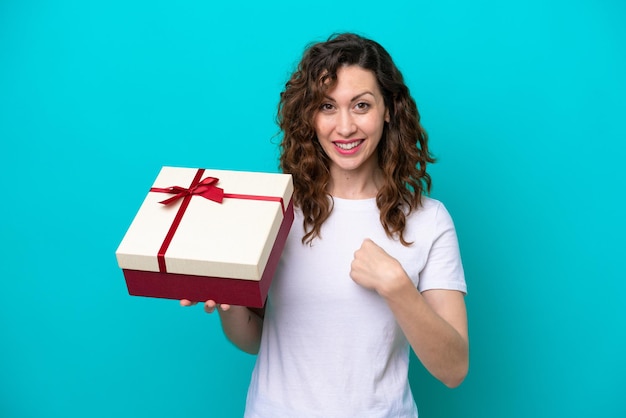 Jeune femme caucasienne tenant un cadeau isolé sur fond bleu avec une expression faciale surprise