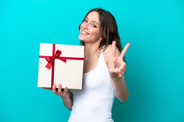 Jeune femme caucasienne tenant un cadeau isolé sur fond bleu souriant et montrant le signe de la victoire