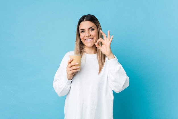 Jeune femme caucasienne tenant un café à emporter joyeux et confiant montrant un geste correct.