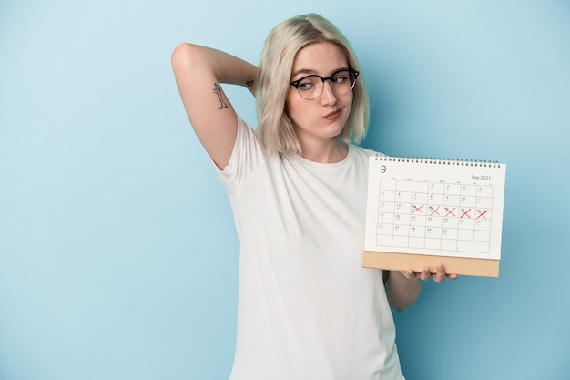 Jeune femme caucasienne tenant un calendrier isolé sur fond bleu touchant l'arrière de la tête, pensant et faisant un choix.