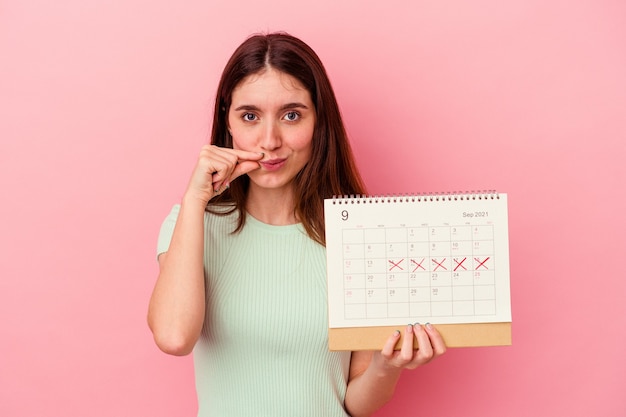 Jeune femme caucasienne tenant un calendrier isolé sur fond rose avec les doigts sur les lèvres gardant un secret.