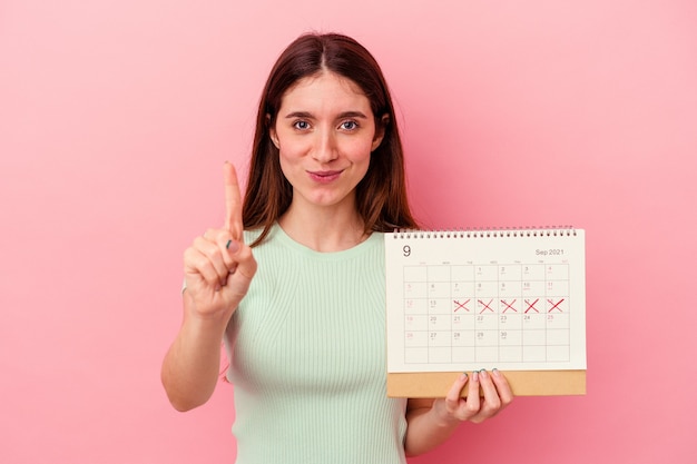 Jeune femme caucasienne tenant un calendrier isolé sur fond rose montrant le numéro un avec le doigt.