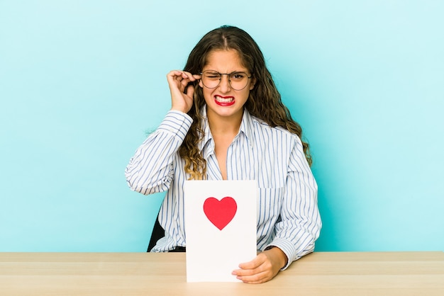 Jeune femme caucasienne tenant une carte de Saint Valentin isolée couvrant les oreilles avec les mains.