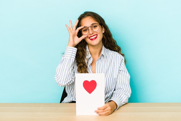 Jeune femme caucasienne tenant une carte de Saint Valentin isolée excité en gardant le geste ok sur les yeux.