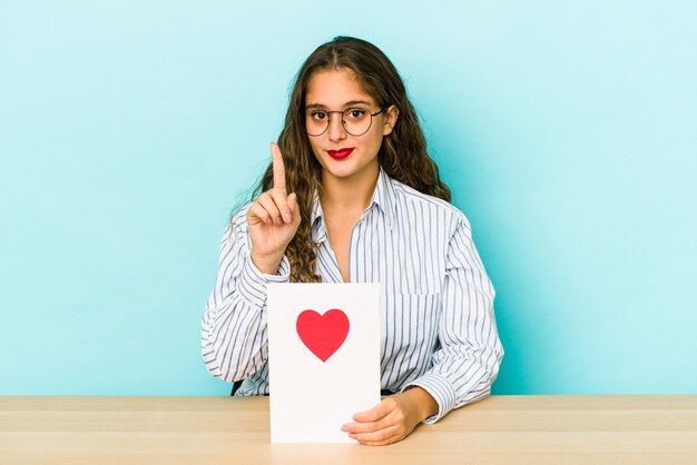 Jeune femme caucasienne tenant une carte de Saint Valentin isolée montrant le numéro un avec le doigt.