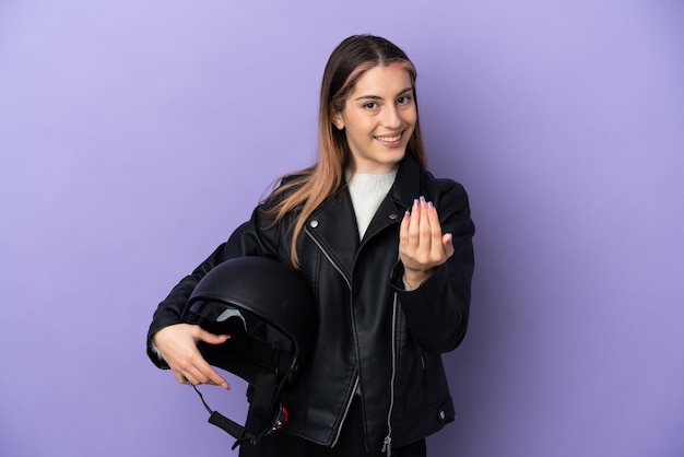 Jeune femme caucasienne tenant un casque de moto isolé sur fond violet invitant à venir avec la main. Heureux que tu sois venu