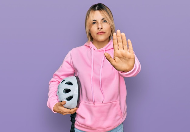 Jeune femme caucasienne tenant un casque de vélo avec la main ouverte faisant un panneau d'arrêt avec un geste de défense d'expression sérieux et confiant