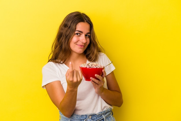 Jeune femme caucasienne tenant des céréales isolées sur fond jaune pointant du doigt vers vous comme si vous vous invitiez à vous rapprocher.