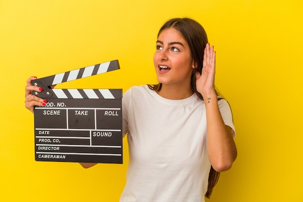 Jeune femme caucasienne tenant un clap isolé sur fond blanc essayant d'écouter un potin.